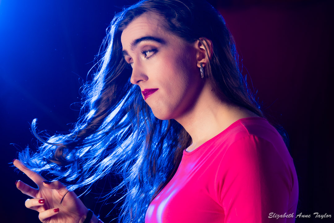 Jasmine's long, brown, wavy hair catches the blue light from off-camera. She wears a pink top, silver and black earrings, red nail polish, and red lipstick. She had her male to female makeover at Elizabeth Taylor's me-to-she dressing service.