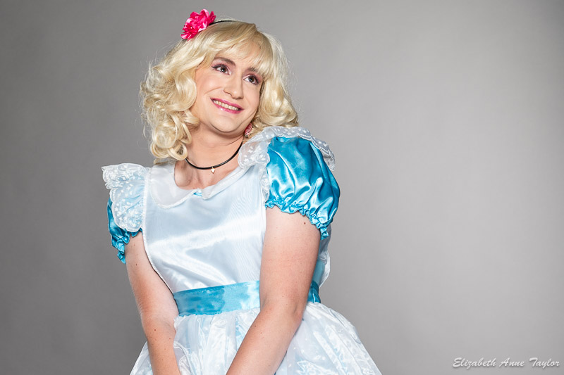A sissy crossdresser wears an Alice in Wonderland dress, black chocker, blonde wig, and a headband with a pink flower on it. She looks upward in expection. She wears pink eyeshadow, pink lipstick, headband, and a blonde wig from Elizabeth Taylor's male to female makeover service.