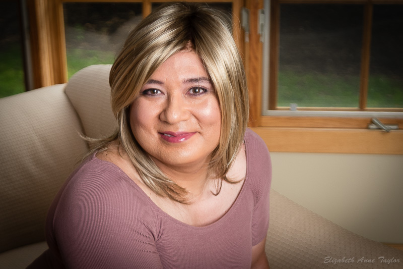 Gina smiles radiantly. She is wearing blonde hair with dark roots and a mauve dress. She's sitting on a chaise lounge and looking up toward the camera. She had an MtF makeover with Elizabeth Taylor in Maryland.