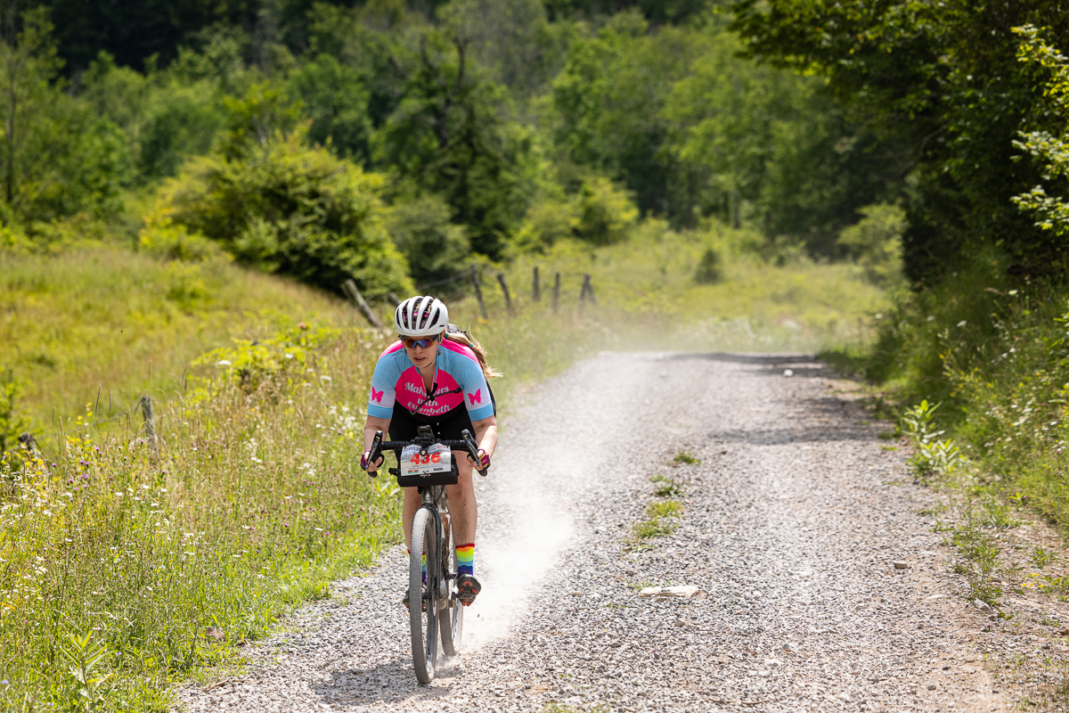 Elizabeth Taylor, owner of Makeovers with Elizabeth Taylor near Washington, DC, flies down a gravel road during GRUSK 2025 in West Virginia wearing her custom pink, blue, and white team kit with the studio name and rainbow socks, race number 436 on her handlebars and a dust cloud trailing behind her. Photographer Bruce Buckley. Used with permission.