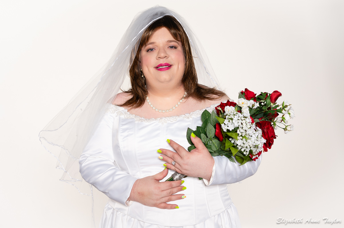 Sissy Bride Mandy wears a white, off-the-shoulder wedding gown complete with a tulle veil while holding her wedding bouquet of red roses. She married her eternal femininity at a sissy bride ceremony at Makeovers with Elizabeth Taylor near Washington, DC.